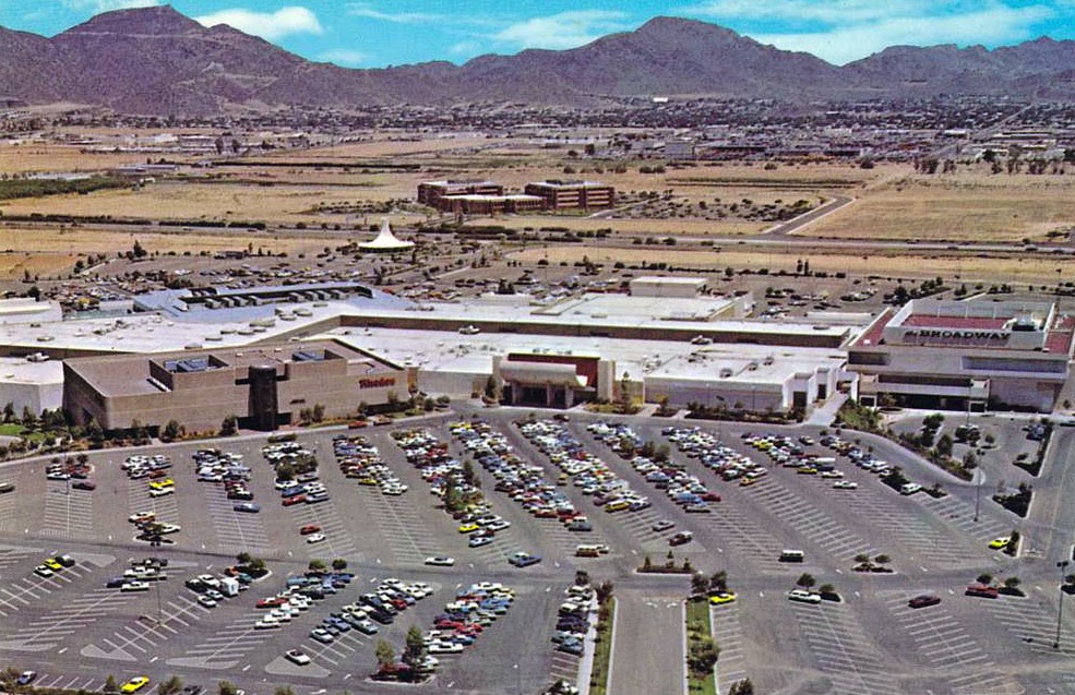 Aerial view of a shopping mall with a large parking lot filled with cars in the foreground. The mall buildings include stores with Safeway visible on the right. In the background are mountains and a desert landscape with scattered development.
