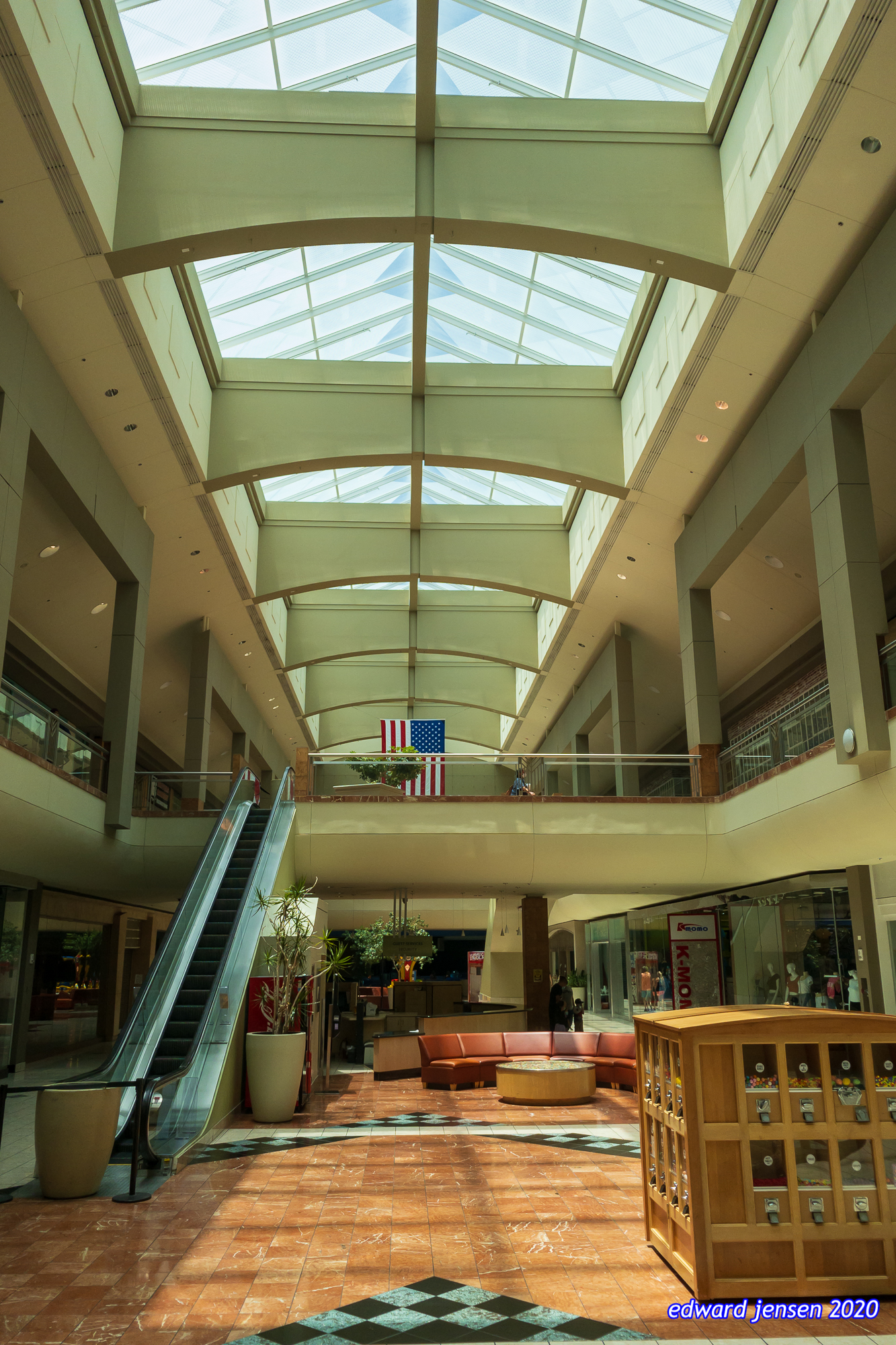 Interior of a shopping mall with a glass ceiling skylight running the length of the building, an American flag hanging from the second floor balcony, escalators on the left, terracotta tiled floor with inlaid patterns, and a seating area with curved orange/pink couches and a round center table.