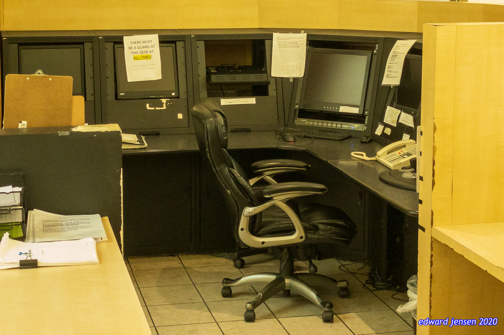 A security desk with multiple computer monitors and a sign that reads "THERE MUST BE A GUARD AT THIS DESK AT ALL TIMES!" A black leather office chair sits at the desk which has a phone and various papers. The desk area has a yellow wall background.