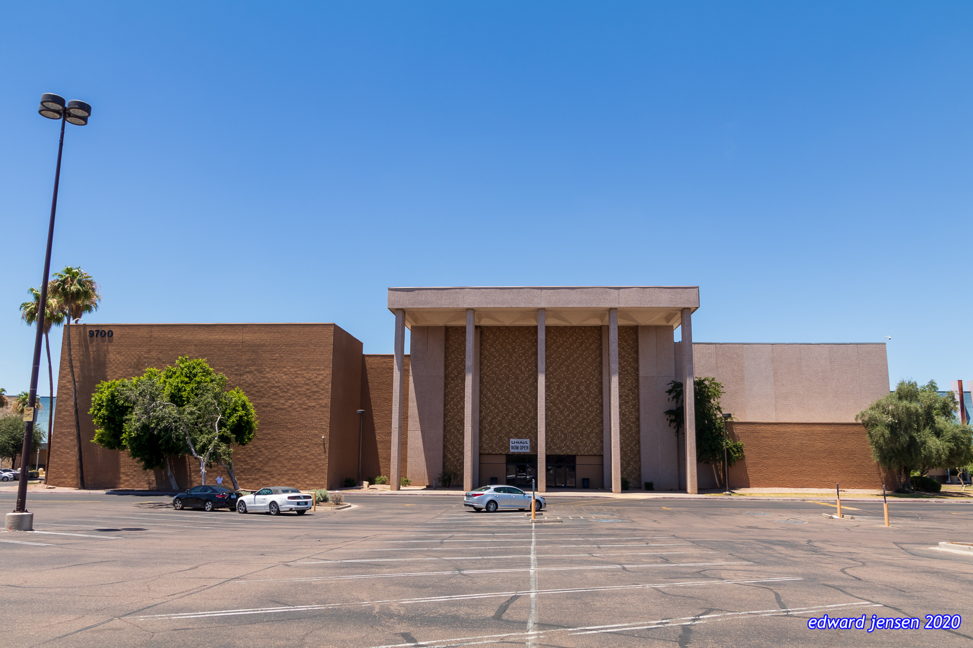 A large mid-century style brick building with a prominent entrance featuring tall columns. The structure appears to be a commercial or institutional building with the number "8700" visible on the left side. Several cars are parked in a large parking lot in front. Palm trees and other landscaping surround the building. The sky is clear blue.