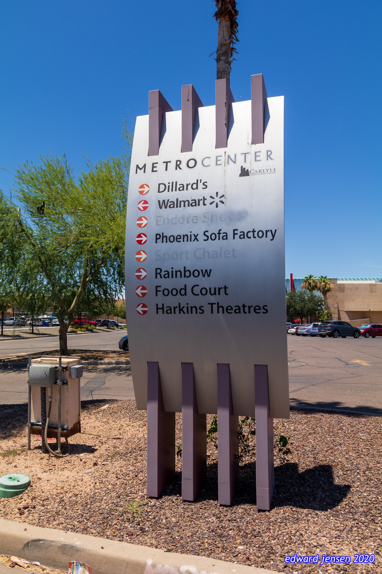 Shopping center sign for MetroCenter with directional arrows pointing to stores including Dillard's, Walmart, Phoenix Sofa Factory, Rainbow, Food Court, and Harkins Theatres, set against blue sky with palm trees and parking lot visible.