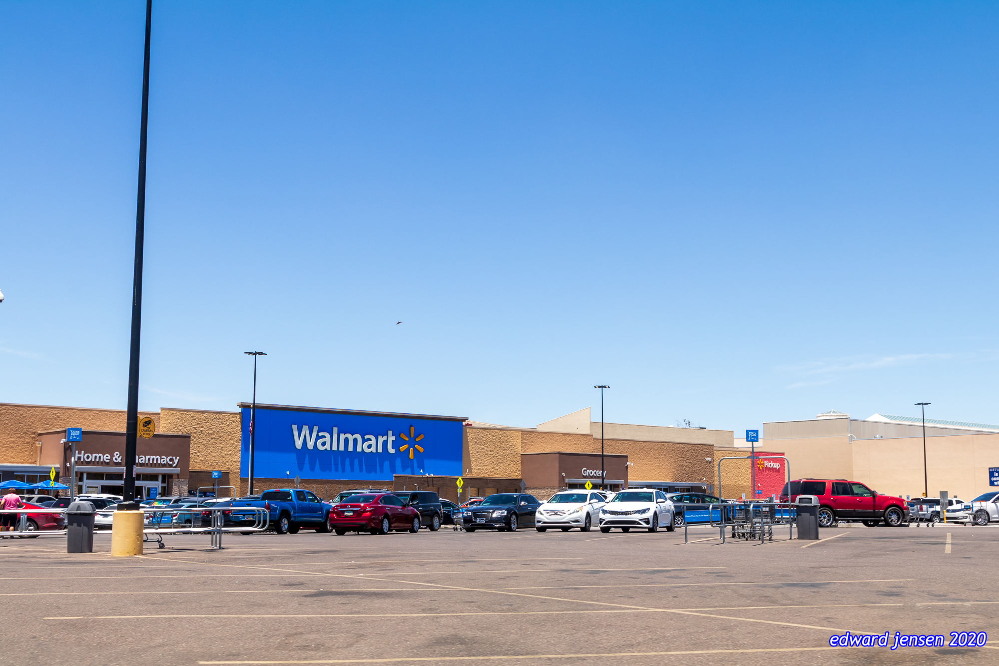 Exterior of a Walmart store with its blue sign and yellow sunburst logo. The building has sections labeled Home & Pharmacy and Grocery. Parking lot in foreground contains various vehicles and shopping cart return areas. Clear blue sky above.
