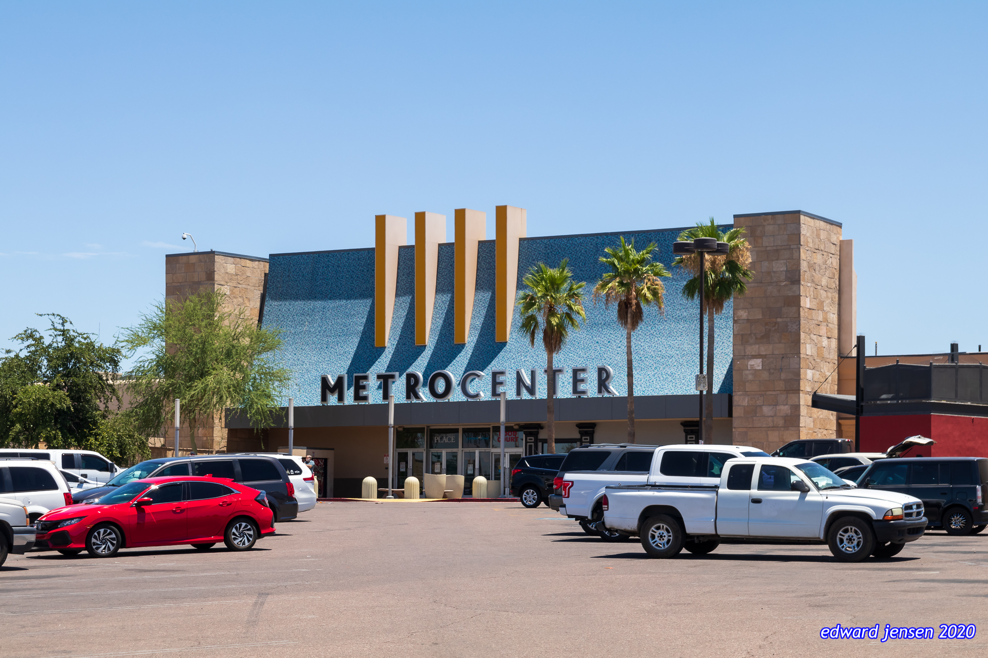 Exterior of a shopping mall called METROCENTER with a distinctive blue facade, vertical gold/tan pillars, and stone accents. Several palm trees stand in front of the entrance. A parking lot with various vehicles including a red car and white truck is visible in the foreground. Clear blue sky overhead.