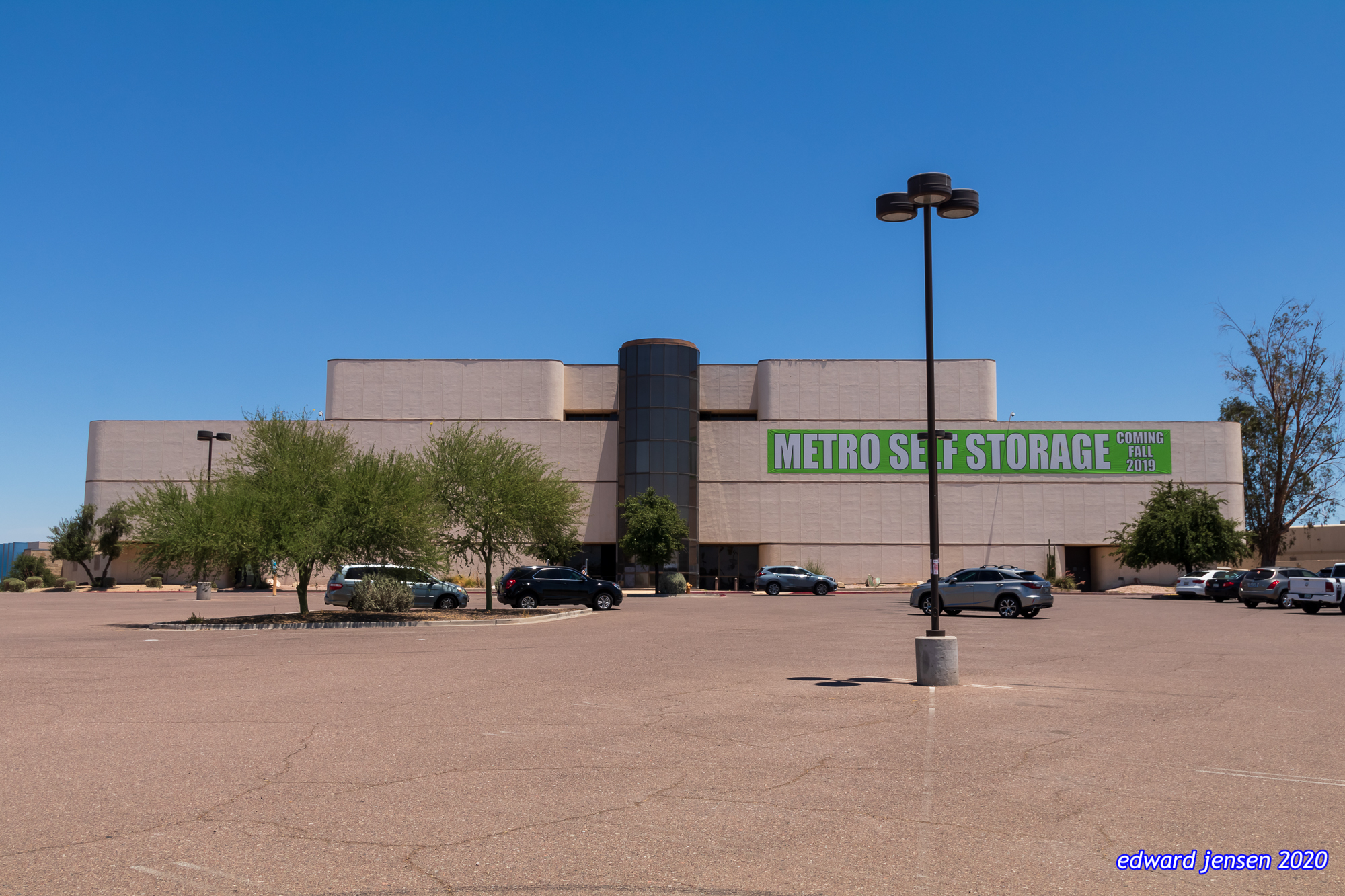 Large beige commercial building with "METRO SELF STORAGE COMING FALL 2019" signage in green letters. The building has a parking lot with several vehicles, desert landscaping with small trees, and a light pole. Clear blue sky background.