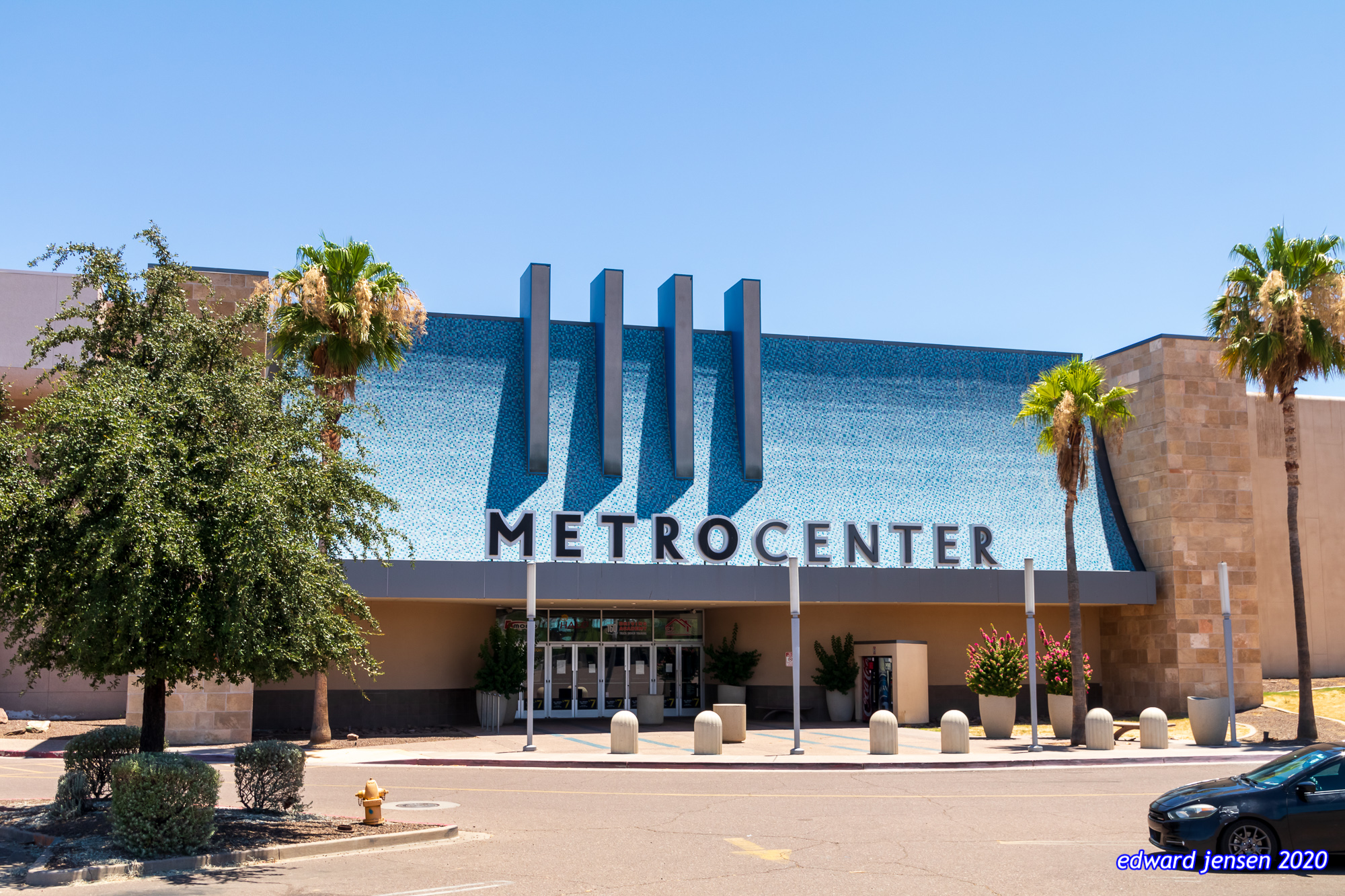 Modern shopping mall exterior with blue facade featuring "METROCENTER" signage, palm trees flanking the entrance, and decorative bollards in the foreground.