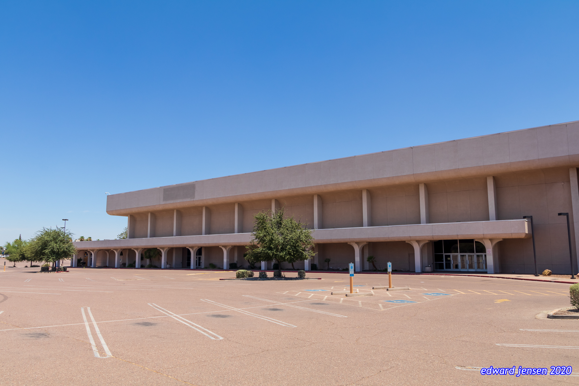 A large beige concrete building with a flat roof and columned entryway, a large anchor store to a former shopping mall. Empty parking lot in foreground with marked spaces including handicap spots. Clear blue sky above.