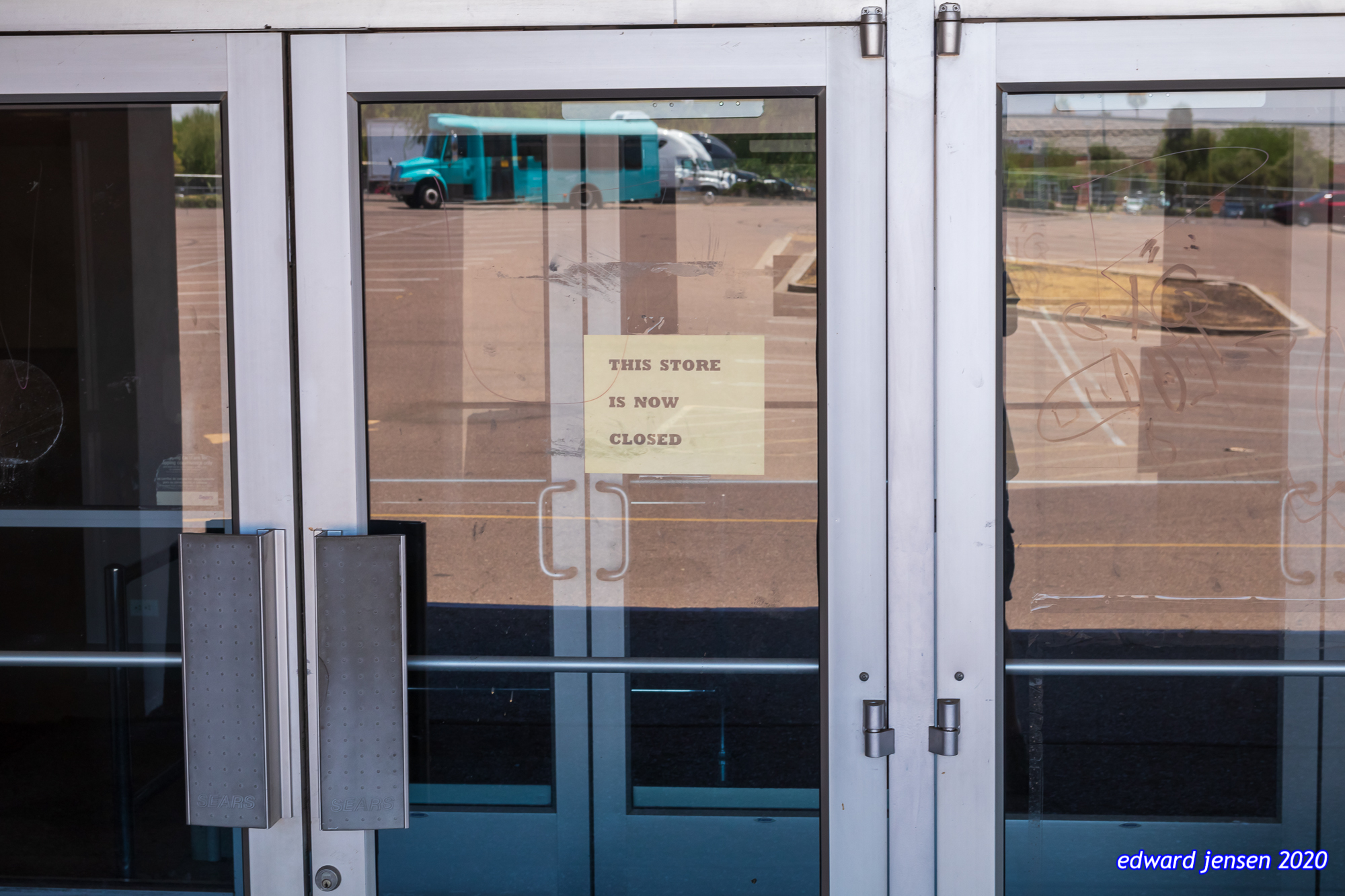 Glass storefront doors with a yellow sign that reads "THIS STORE IS NOW CLOSED". A turquoise bus is reflected in the glass.