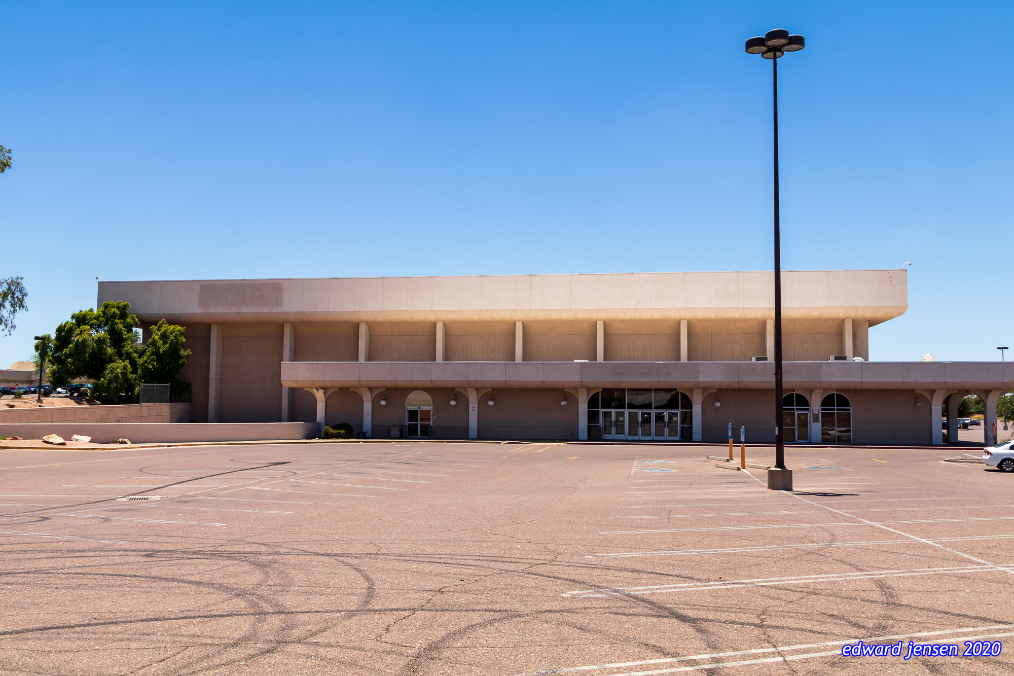 A large beige commercial building with a flat roof and columns at the entrance, viewed from across a mostly empty parking lot. The building has a modernist design with a tall light pole in the foreground. Trees are visible on the left side. Clear blue sky above.
