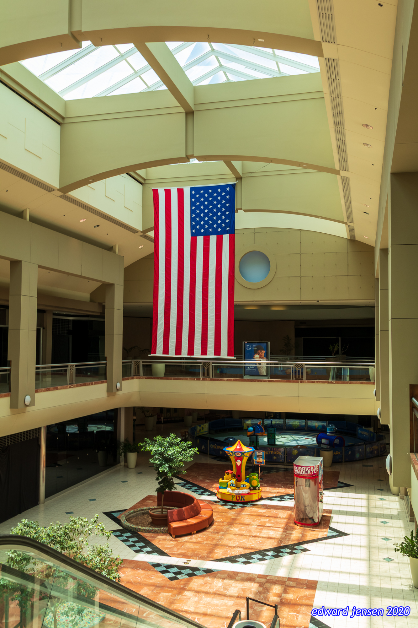 Interior of a shopping mall with a large American flag hanging from the ceiling. The mall features skylights, a two-level layout, and a central courtyard area with children's rides marked "FUN" and a "HURRICANE SIMULATOR" attraction. Small trees and plants decorate the space.