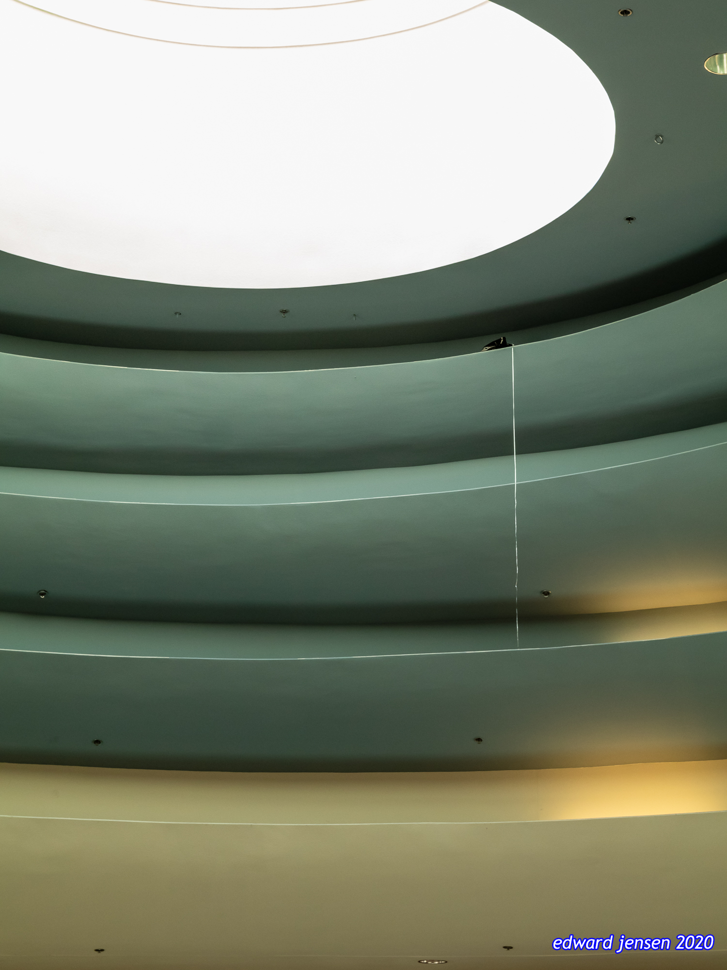 Modern architectural interior with curved ceiling layers in pale green tones transitioning to cream at the bottom, with a circular skylight at the top. The ribbon from a balloon caught in one of the ceiling layers hangs from the ceiling.