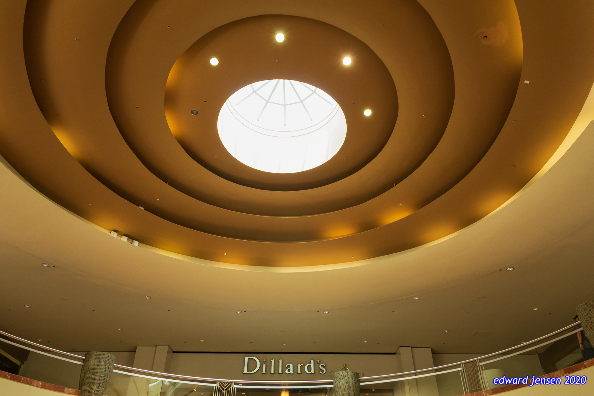 Architectural interior showing a circular gold ceiling with concentric rings, featuring a bright central skylight dome. Below is the entrance to Dillard's department store with illuminated signage.