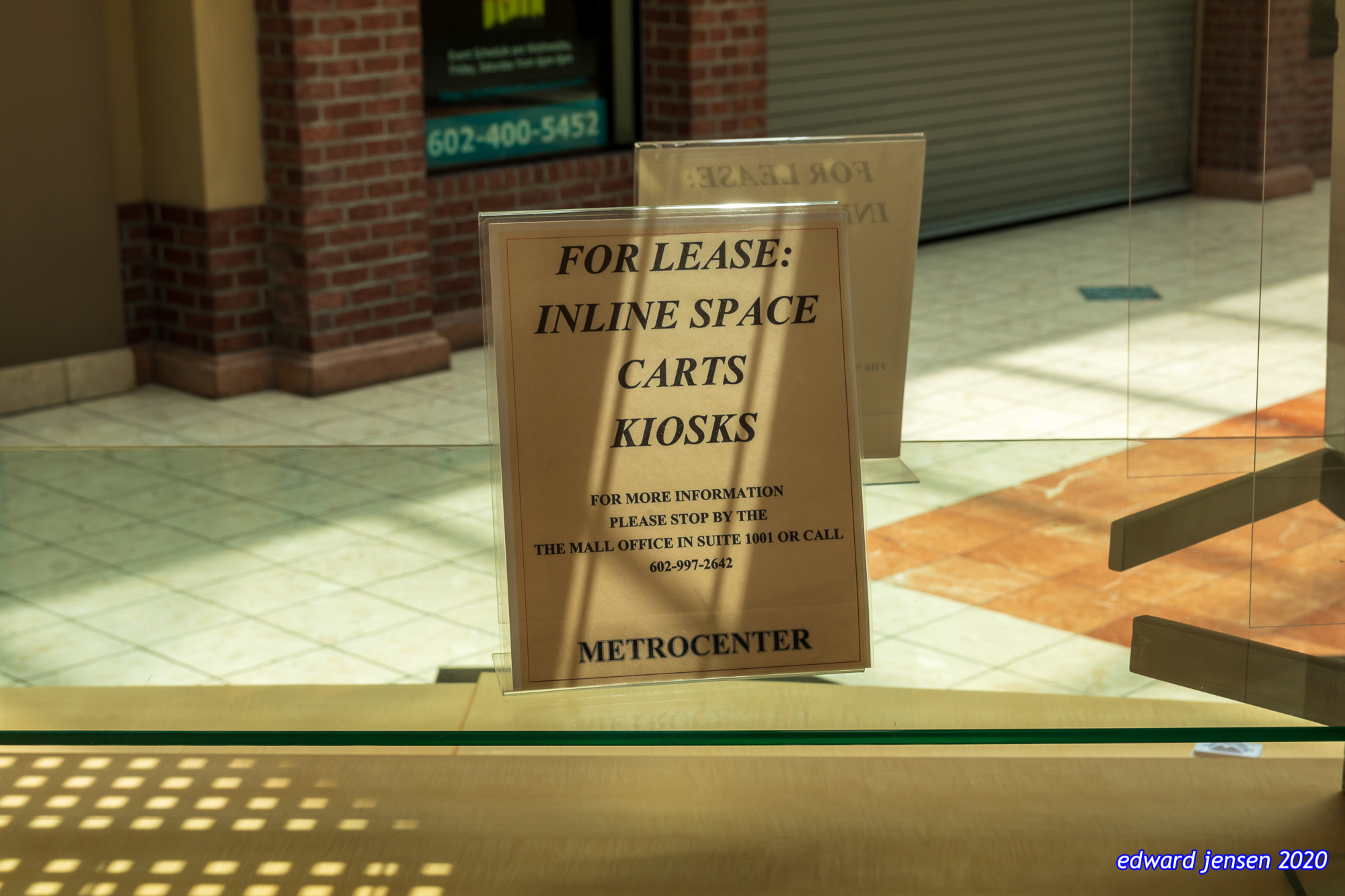 Sign in a shopping mall: FOR LEASE: INLINE SPACE CARTS KIOSKS FOR MORE INFORMATION PLEASE STOP BY THE THE MALL OFFICE IN SUITE 1001 OR CALL [phone number redacted] The phone number is visible in the reflection of a closed store window.