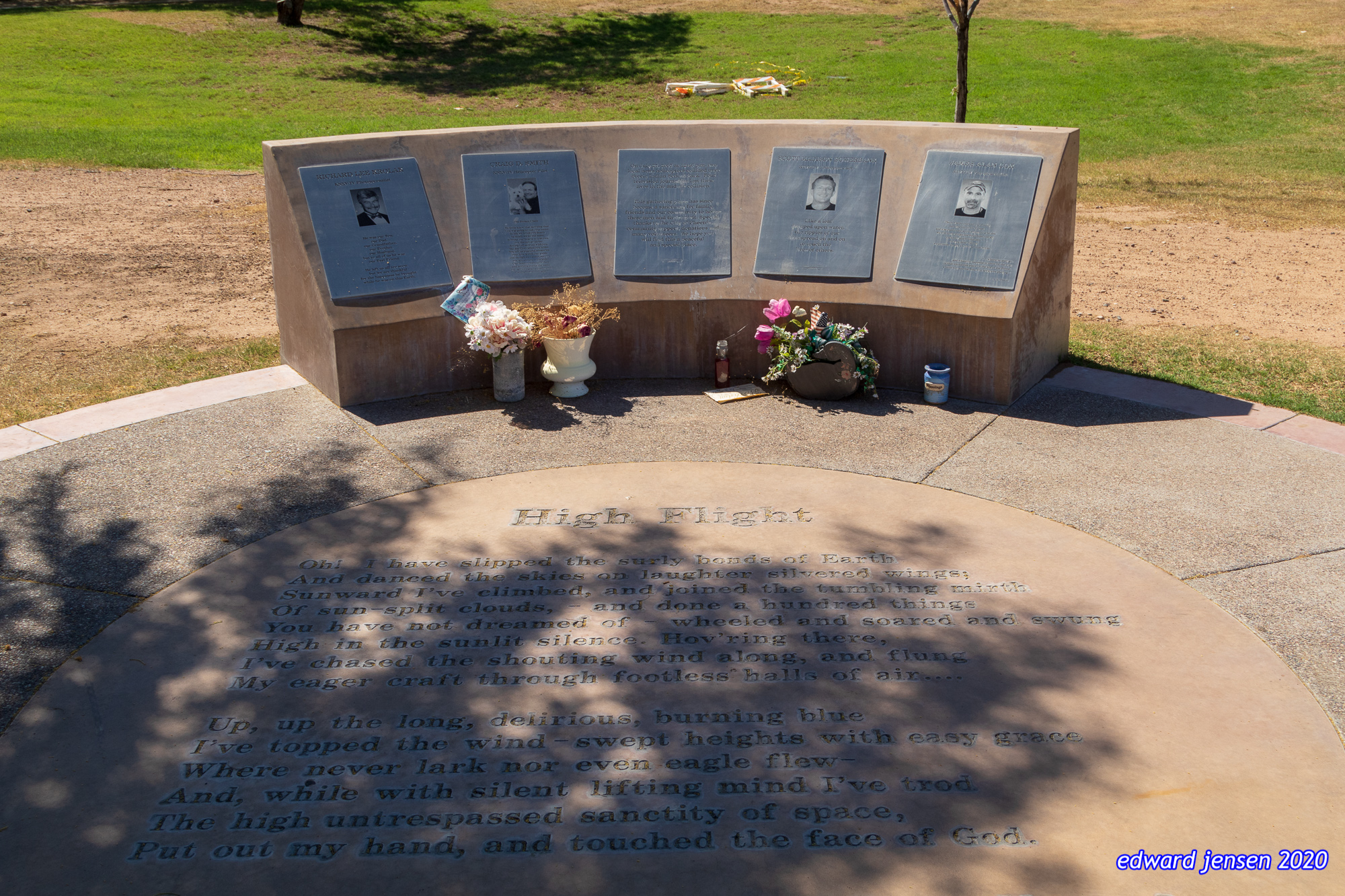 A memorial site with a curved stone wall containing five plaques with photographs, and a circular ground inscription titled "High Flight" containing a poem about flight that begins "Oh! I have slipped the surly bonds of Earth" and ends "Put out my hand, and touched the face of God." Flower arrangements are placed at the base of the memorial.