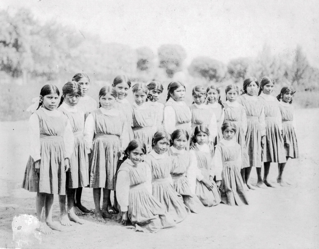 Historical black and white photograph showing a group of Native American girls in two rows wearing similar light-colored dresses, standing outdoors on bare ground with trees in the background.