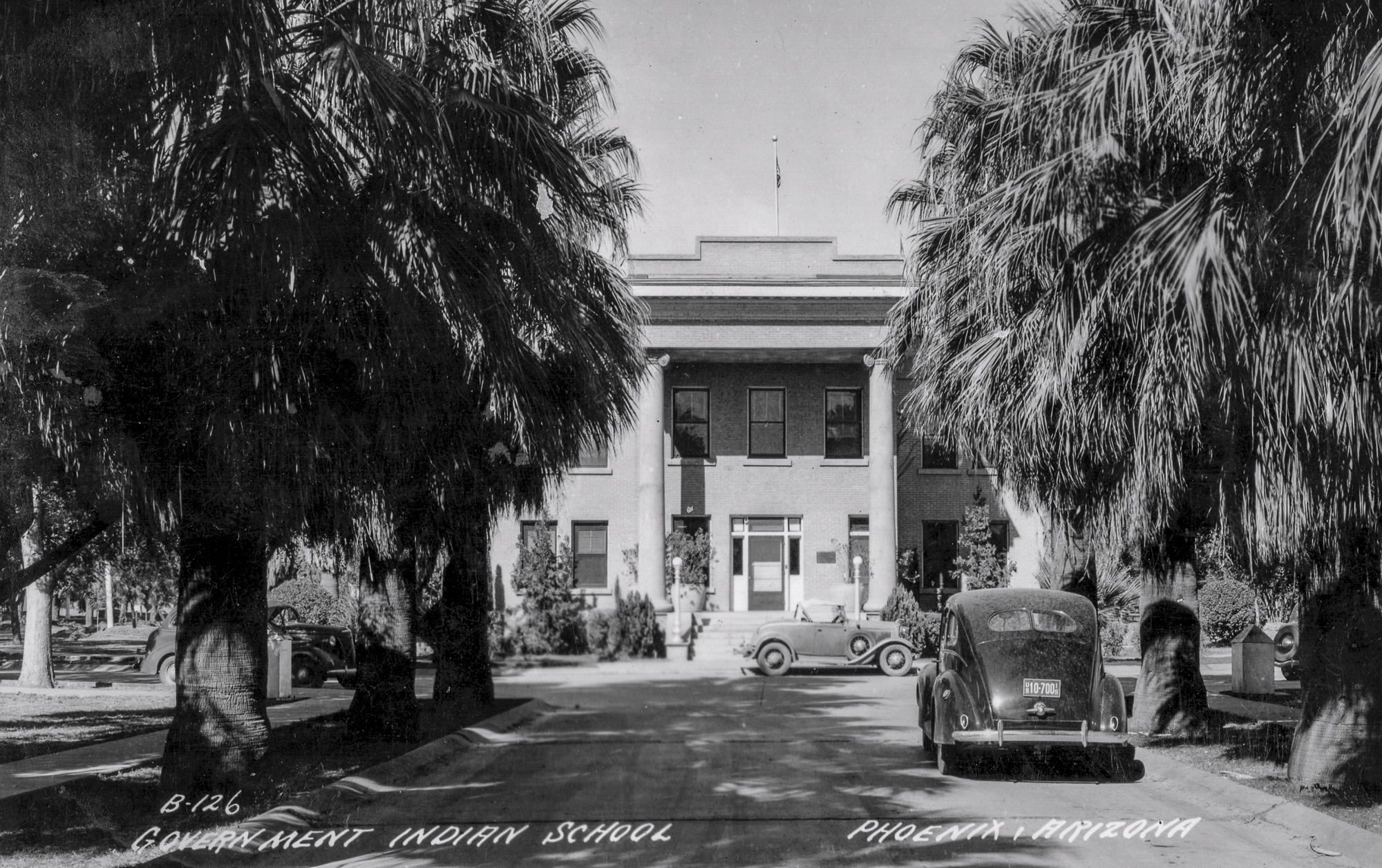 Historic black and white photograph of Government Indian School in Phoenix, Arizona. A columned building with palm trees on either side of the entrance path. Vintage cars are parked in front. Text at bottom reads: B-126 GOVERNMENT INDIAN SCHOOL PHOENIX, ARIZONA