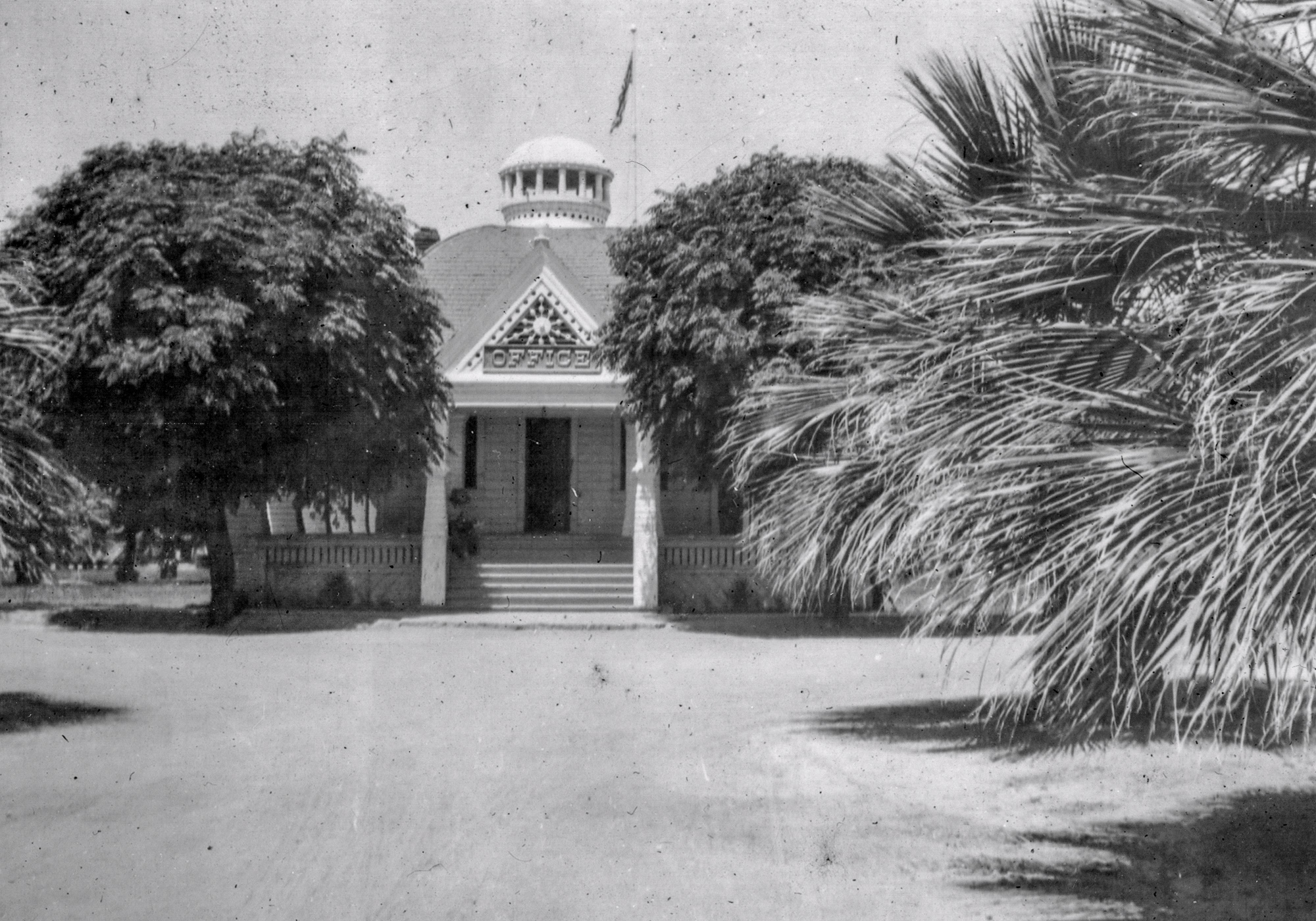 Historical black and white photograph of a colonial-style building with a domed cupola and flag on top. The building features ornate triangular decorative elements above its entrance. It is flanked by large trees on the left and palm fronds visible on the right, creating a symmetrical approach to the front steps.