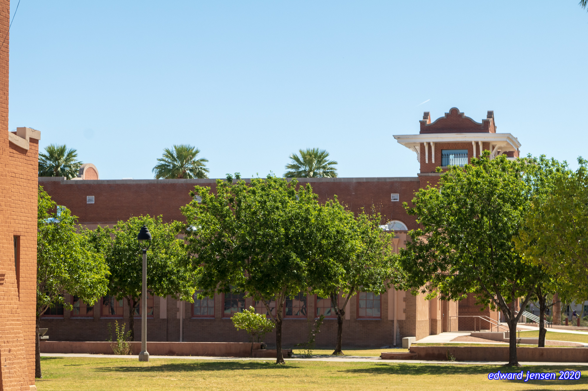 Historic brick building with architectural details, surrounded by lush green trees and palm trees visible above a brick wall. A decorative tower or cupola structure is visible on the right side. Grassy courtyard area in foreground with a street lamp.