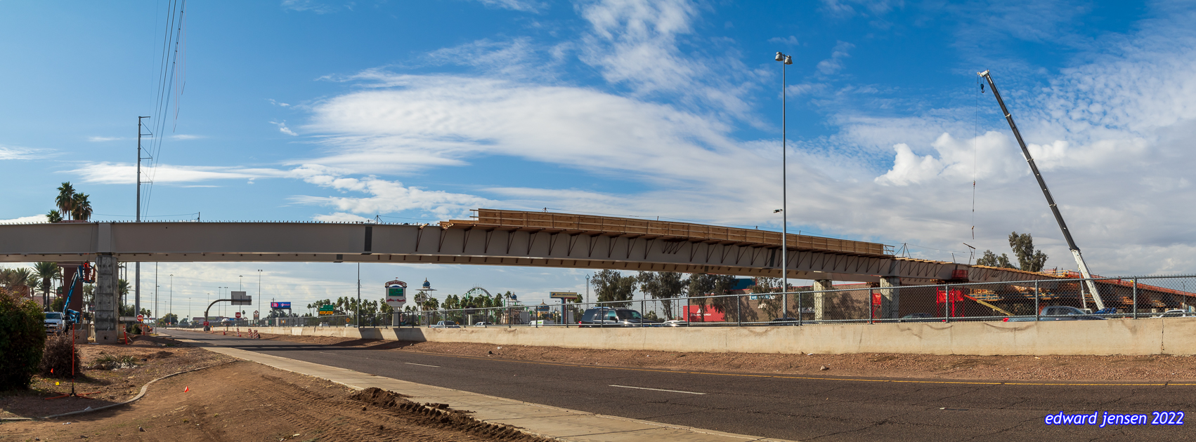 Panoramic view of a highway construction site showing an overpass bridge being built, with a wooden formwork structure supported by concrete pillars. Construction equipment including a crane is visible at the site, surrounded by safety barriers. Palm trees line the background under a bright blue sky with scattered clouds.