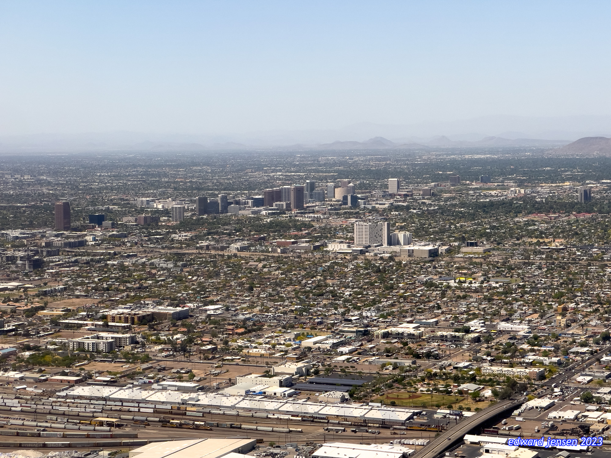Aerial view of Phoenix, Arizona cityscape showing downtown skyscrapers surrounded by urban sprawl, with mountains visible in the background and train yards in the foreground.