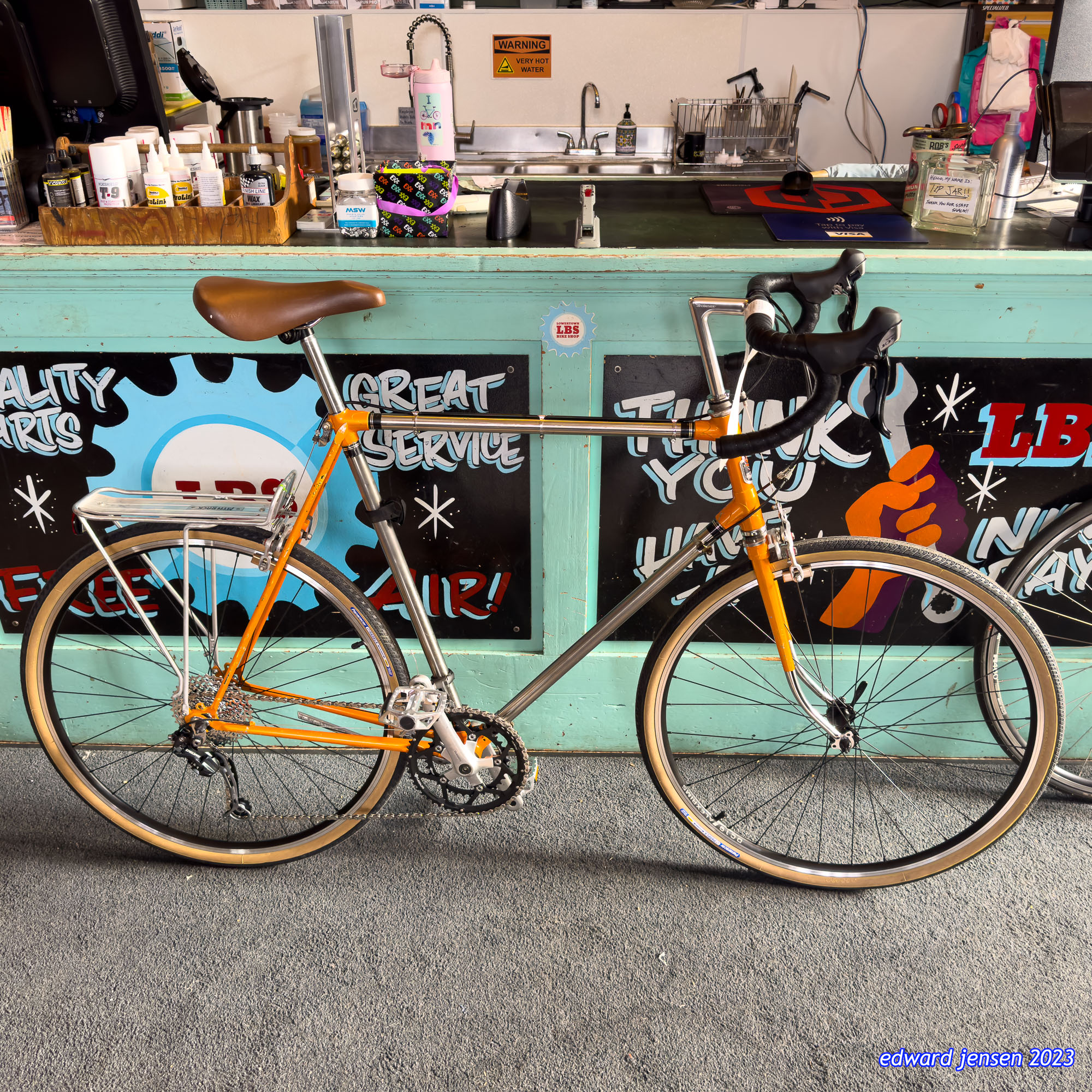 An orange and silver road bike with drop handlebars and brown leather saddle parked in front of a bike shop counter. Behind it is a mint green counter with colorful signage reading "GREAT SERVICE" and "THANK YOU". Above the counter is a sink area with a "WARNING VERY HOT WATER" sign.