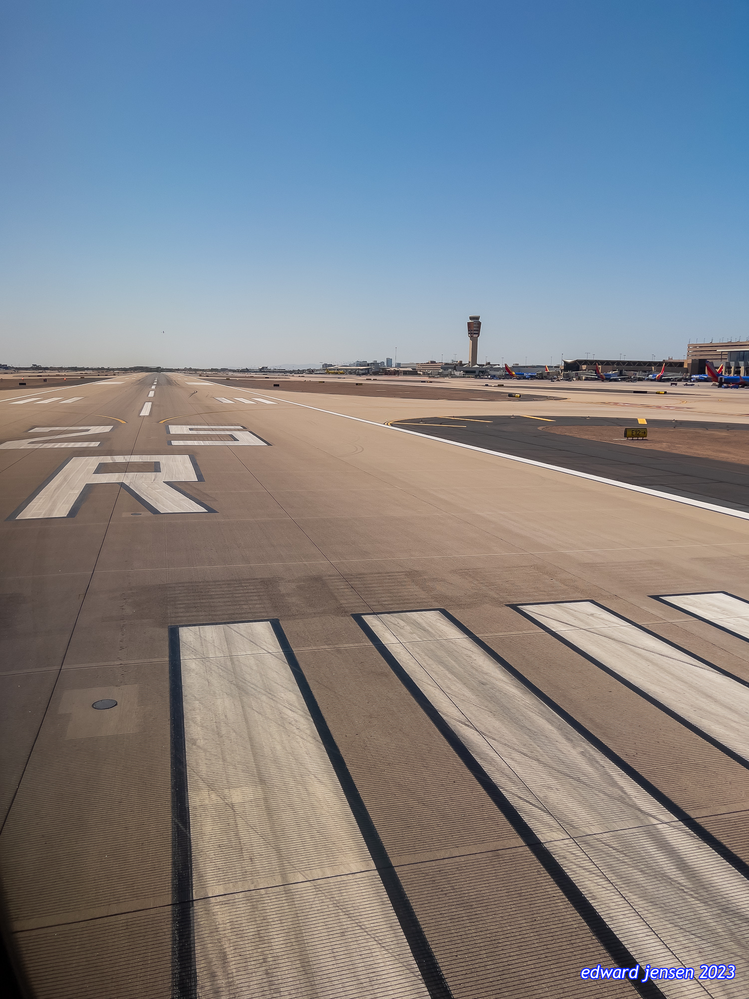 Airport runway view from the tarmac, showing runway markings with large markings 25R, control tower in the distance, parked aircraft at terminal, and clear blue sky.