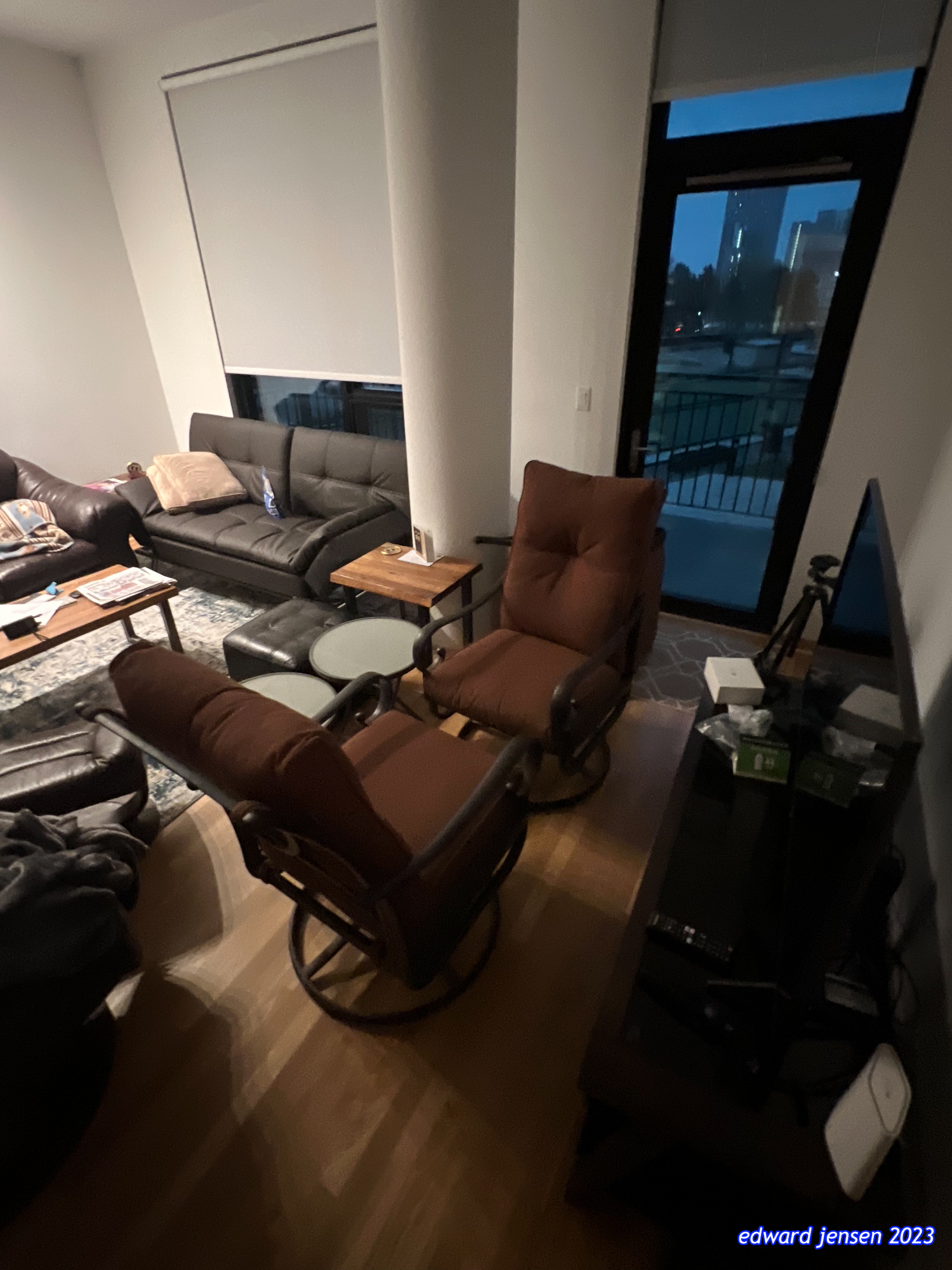 Modern apartment living room with a black leather sofa, two brown reclining chairs, wooden coffee table, and large glass door/window showing city view at dusk. Wood flooring, white walls, and a window shade visible. Small side tables and items on surfaces.
