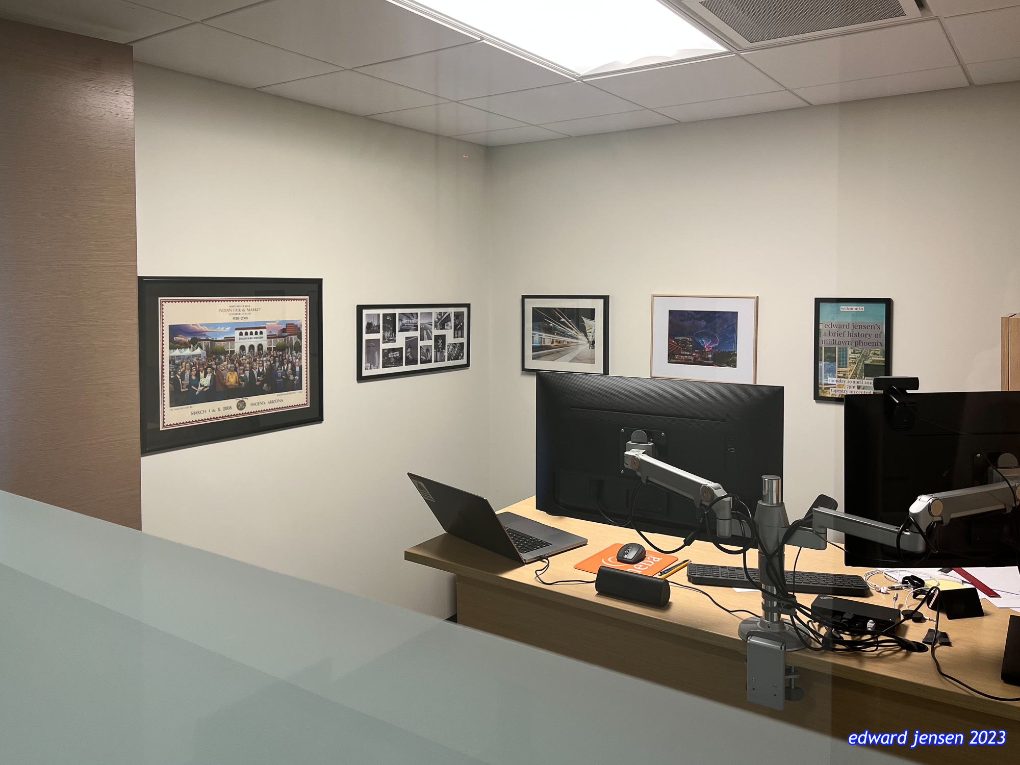 Office workspace with wooden desk featuring a laptop and dual monitor setup on articulating arms. White walls display several framed photographs including one labeled "Edward Jensen's A Brief History of Midtown Phoenix" and what appears to be an official event photo.