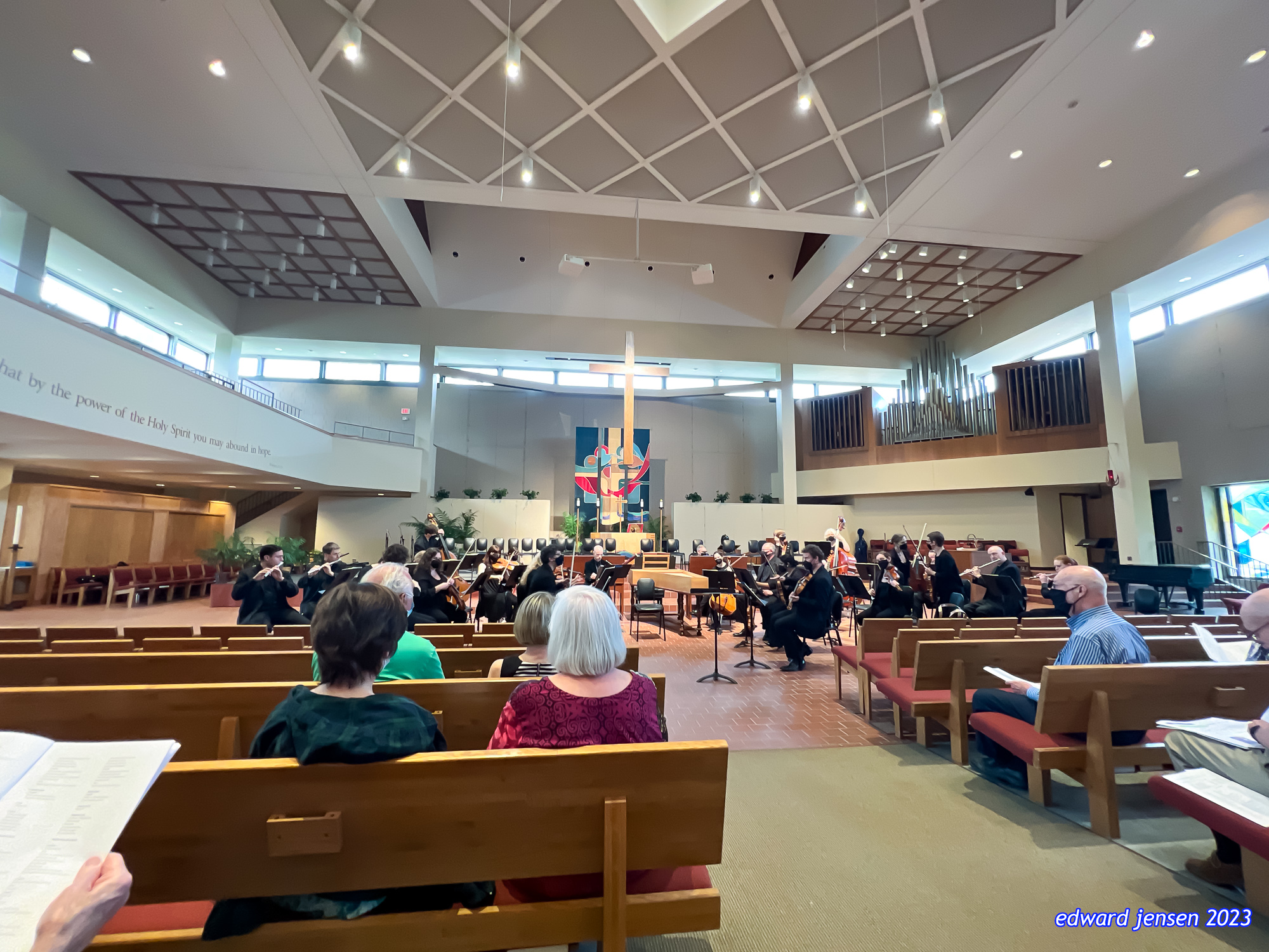 Interior of a church during an orchestra performance. Musicians in formal attire playing instruments on a stage in front of a colorful stained glass or artwork cross design. A pipe organ is visible on the right wall. Audience members seated in wooden pews. The ceiling has a geometric pattern with pendant lights. Partial text visible on wall: "...by the power of the Holy Spirit you may abound in love."