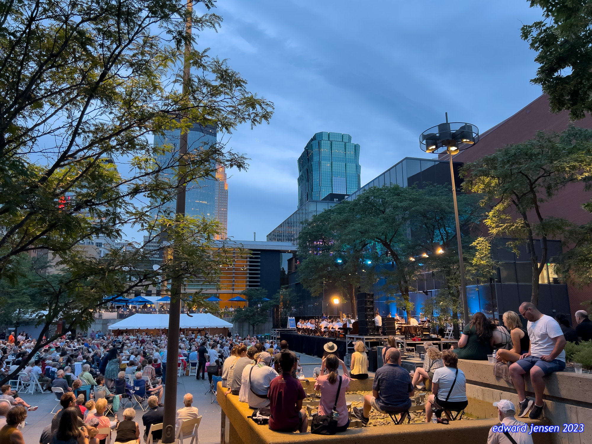 Outdoor evening concert in an urban setting. A large crowd sits in chairs watching a performance on a stage with musicians. The venue is surrounded by tall modern skyscrapers with glass facades and trees. City lights are beginning to shine as dusk settles.