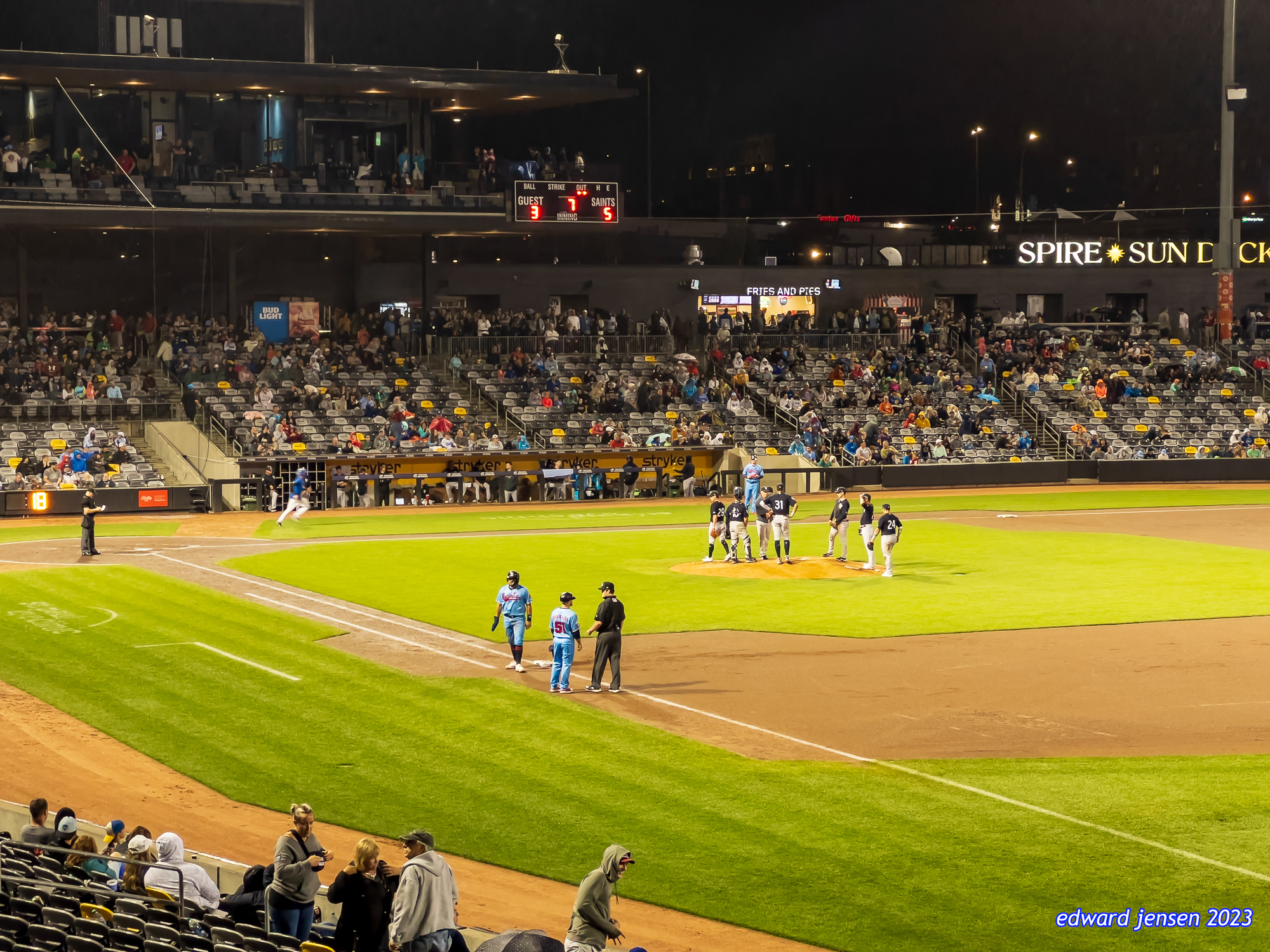 Nighttime baseball game in a stadium showing the field with players in light blue uniforms near third base, a scoreboard displaying GUEST: 3, SAINTS: 5, partially filled stands, and concessions area labeled "FRIES AND PIES".