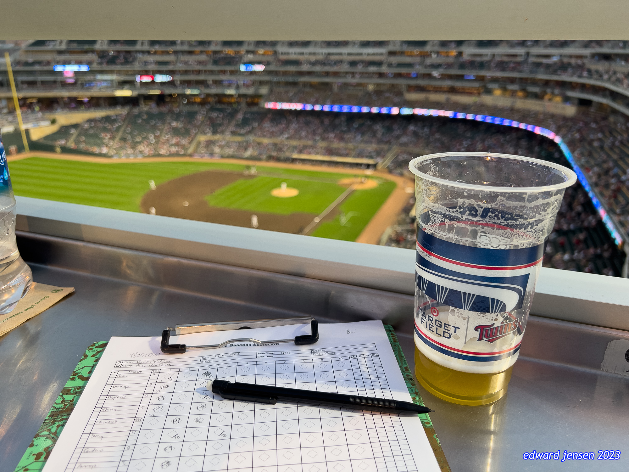 View from stadium seating at Target Field, home of the Minnesota Twins. In the foreground is a baseball scorecard with pencil and a cup of beer featuring the Target Field and Twins logos. The baseball diamond and field are visible below, with stadium seating and lighting visible in the background.