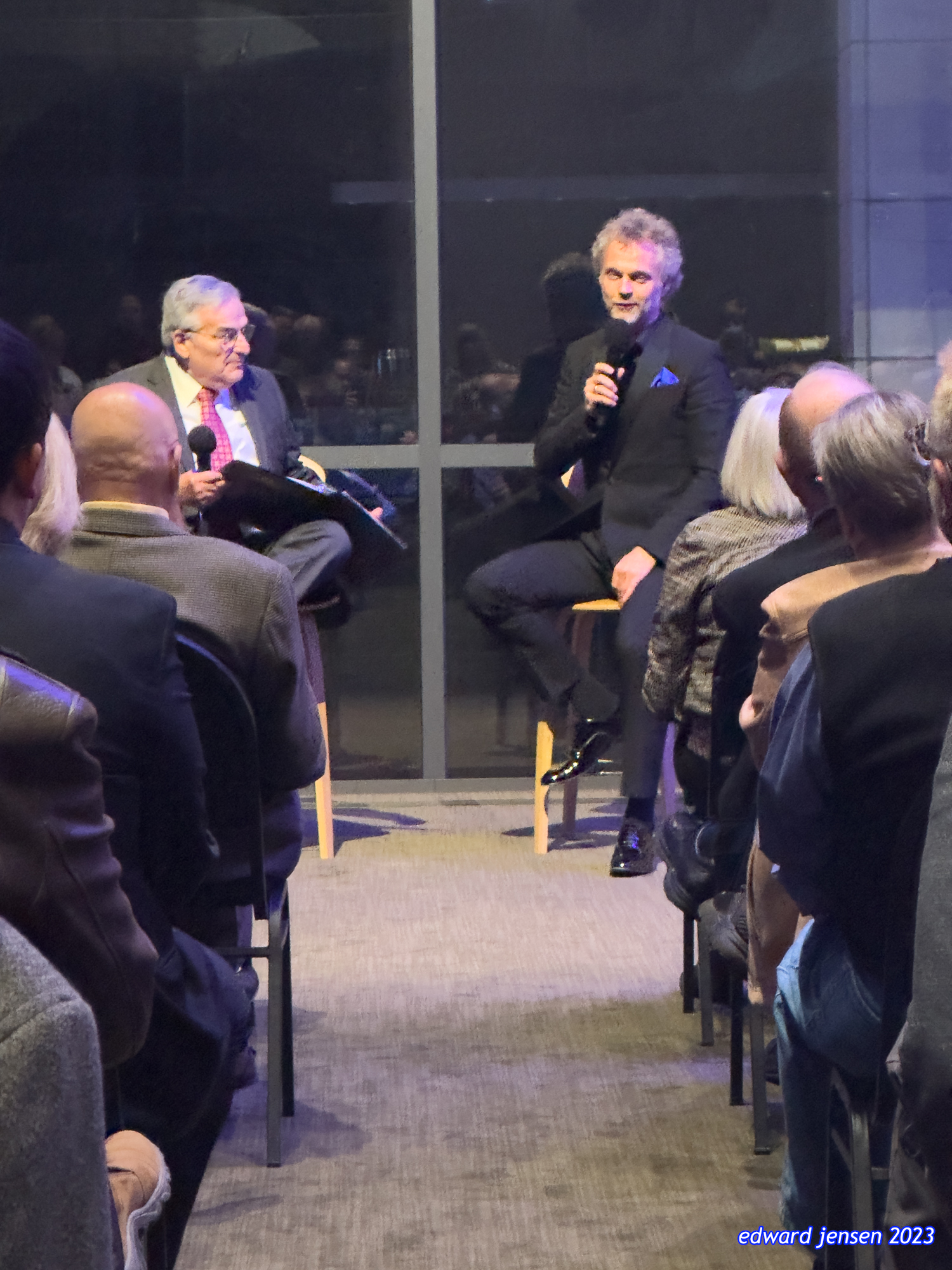A panel discussion or interview in a modern venue. Two speakers on stage holding microphones - one older man in a suit with glasses on the left and another man in a dark suit with a blue pocket square on the right. They're seated on wooden stools facing an audience. The audience is viewed from behind, seated in rows.