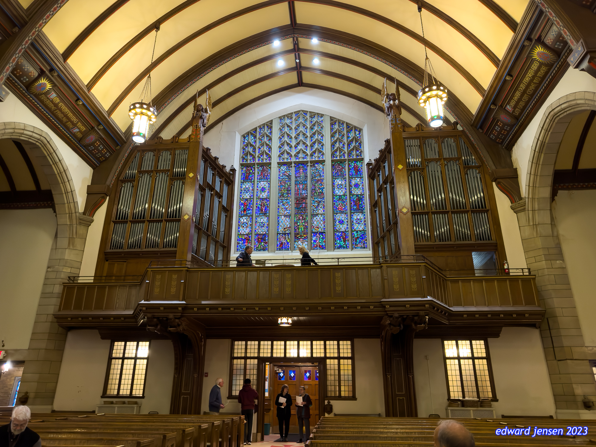 Interior of a church with a magnificent pipe organ and stained glass window. The vaulted ceiling has wooden arches with pendant lights. Two people visible at the organ console beneath the colorful stained glass. Several people standing in the doorway and some seated in pews.