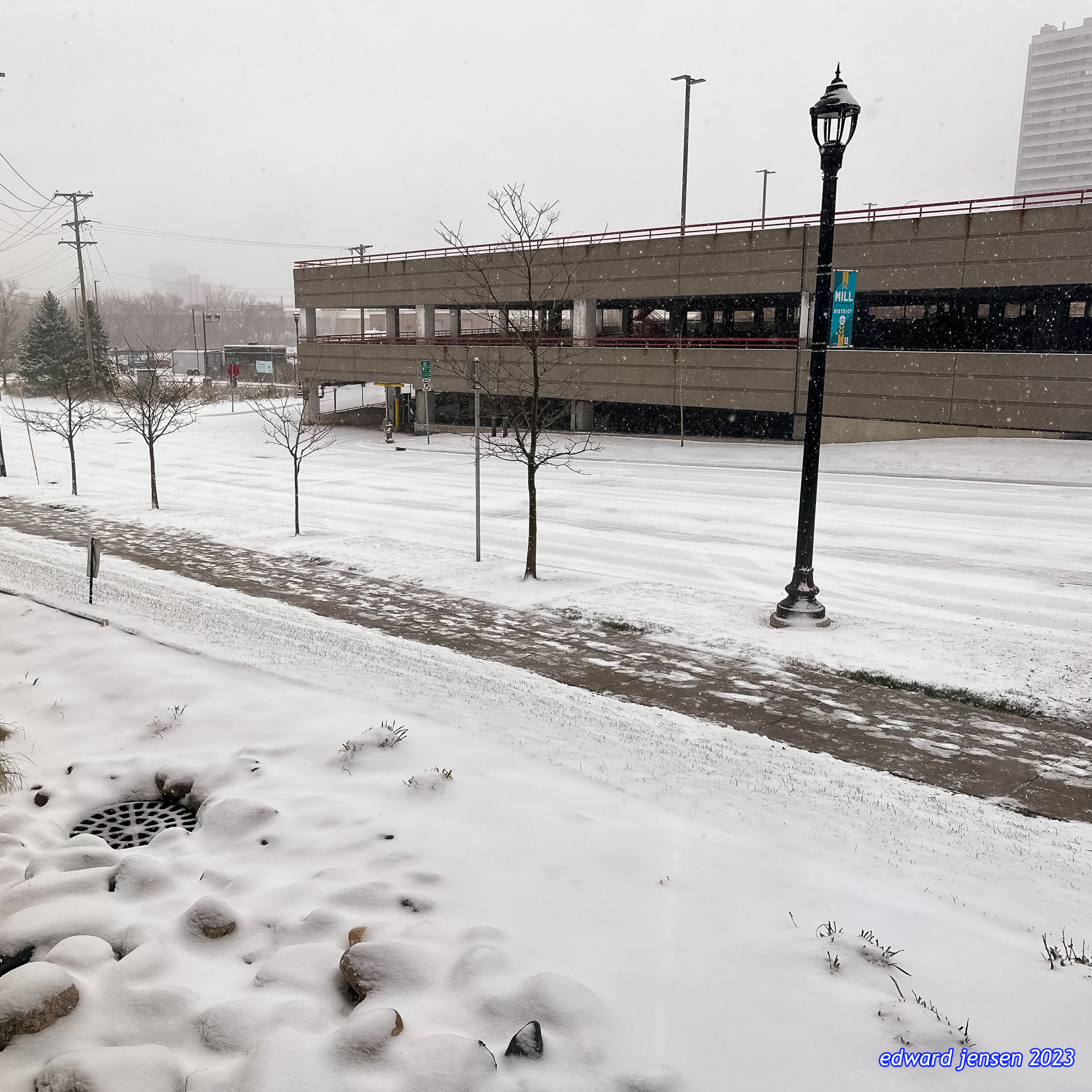 A snowy winter scene showing a parking garage or structure with concrete levels visible, snow-covered ground with partially cleared walkways, several bare trees, a black vintage-style lamppost, and light snowfall. Power lines are visible in the background.
