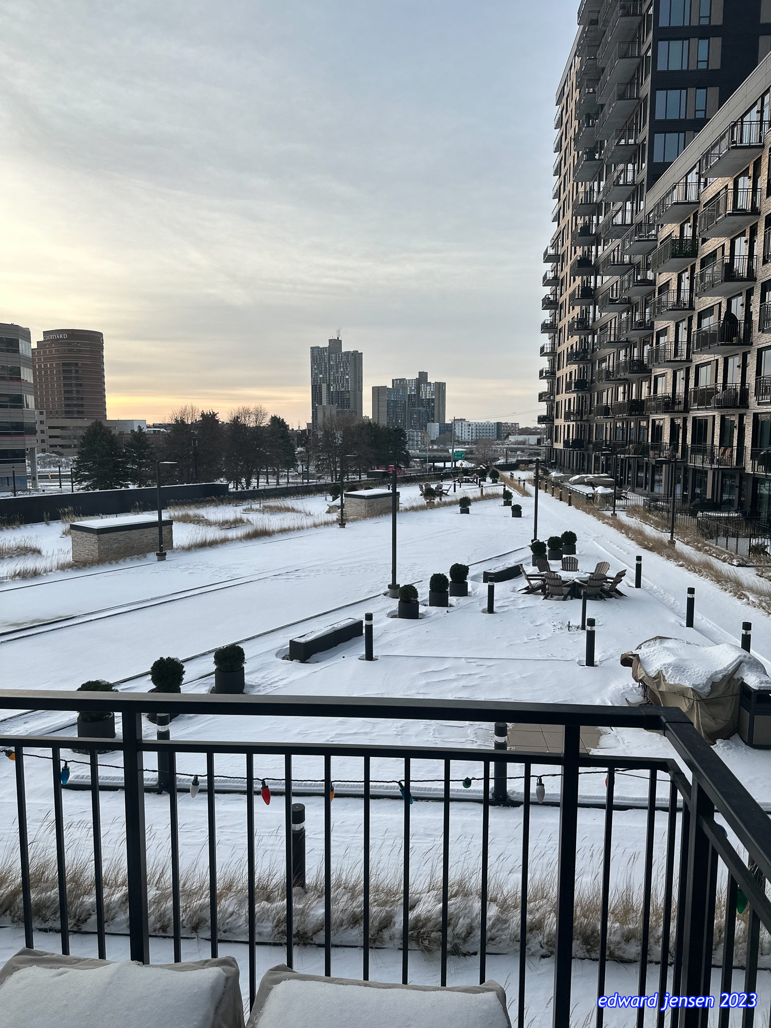 Winter urban landscape view from a balcony overlooking a snow-covered courtyard with benches and outdoor seating area. Tall residential buildings with many balconies visible on the right side, with a city skyline featuring several high-rises in the background. The scene is captured at dusk with a cloudy sky.