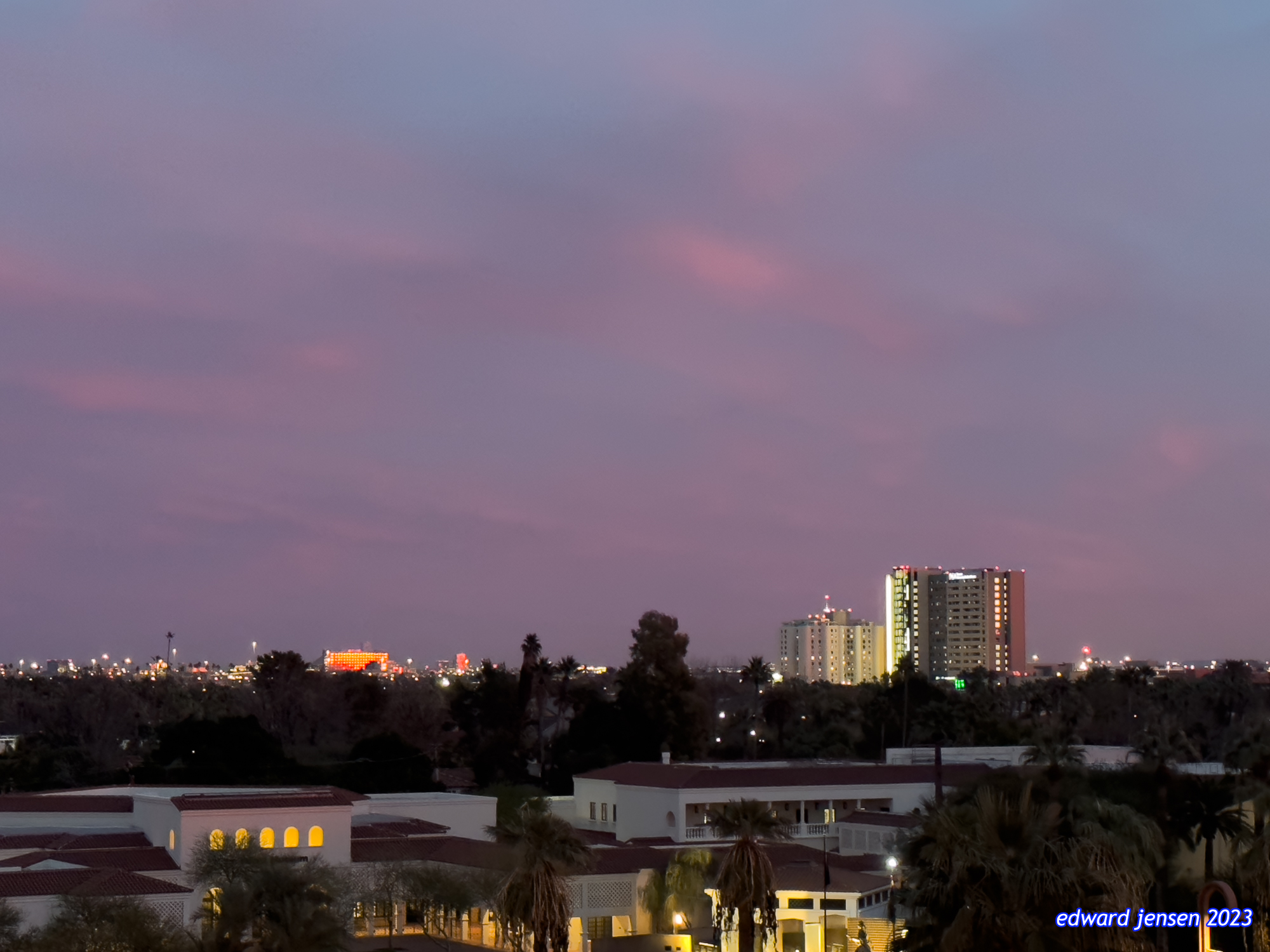 Urban skyline of Phoenix, Arizona, at dusk with purple sky, showing lit buildings, palm trees, and distant high-rises. The foreground shows lower structures with illuminated windows while taller buildings shine in the distance.