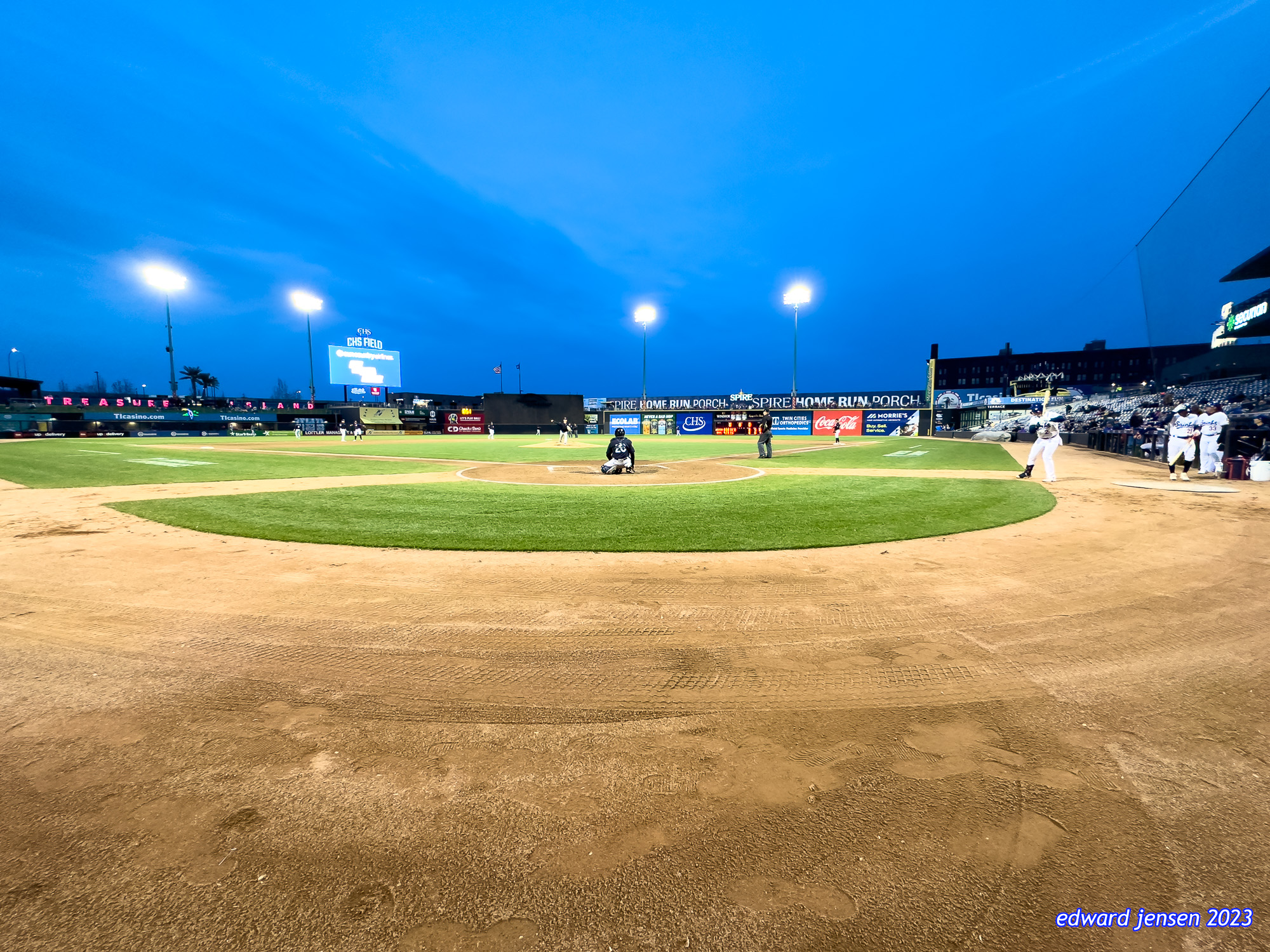 Baseball stadium at twilight showing a minor league field with illuminated field lights against a blue evening sky. Visible advertisements include SPIRE HOME RUN PORCH, Coca-Cola, and TREASURE ISLAND. Players positioned on the field with the pitcher's mound and home plate visible.