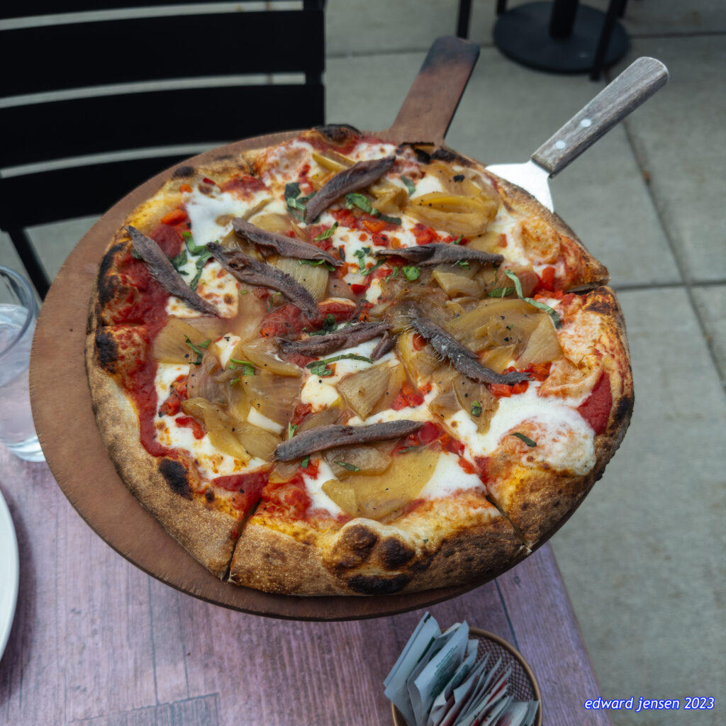 Wood-fired pizza on a wooden board with melted cheese, tomato sauce, caramelized onions, anchovy fillets, and herbs. A server's knife with wooden handle is visible on the pizza.