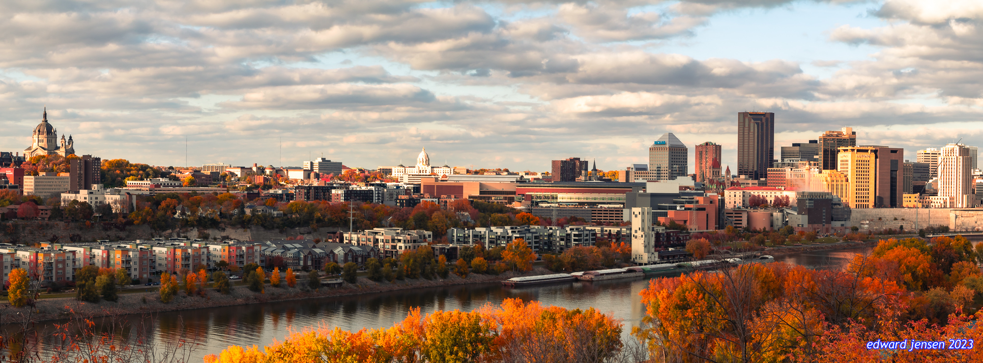 Panoramic cityscape of St. Paul, Minnesota in autumn with colorful fall foliage surrounding the Mississippi River, downtown skyscrapers, and the Cathedral of Saint Paul visible on the left under a cloudy sky.